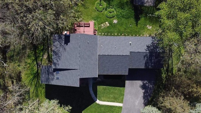 Aerial view of a newly installed dark asphalt shingle roof featuring two rectangular skylights and a brick chimney, surrounded by mature trees and a landscaped backyard with a red wooden deck—showcasing expert craftsmanship, modern design elements, and enhanced energy efficiency in a residential roofing project.